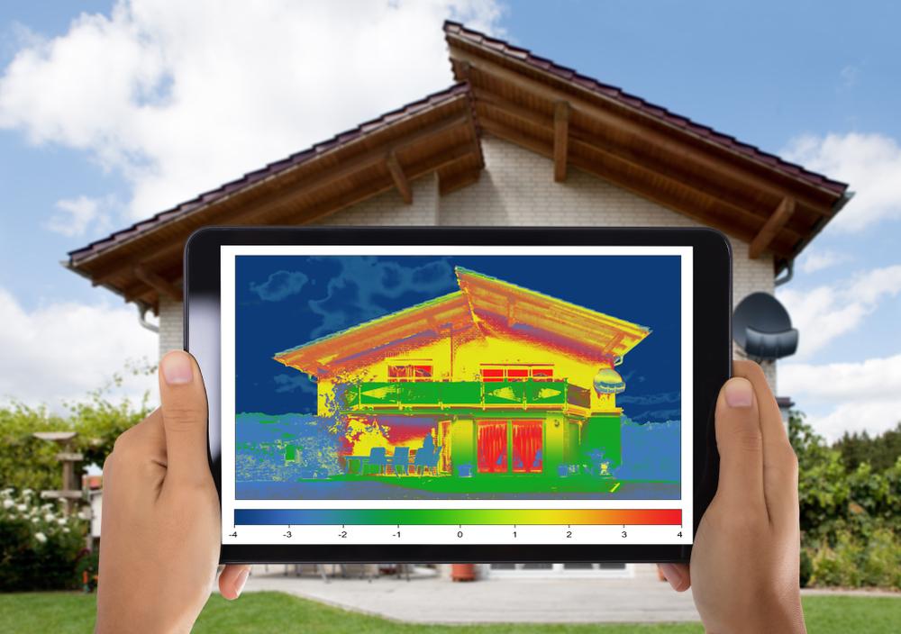 Person holding a tablet displaying a thermal image of a house in front of the corresponding house.